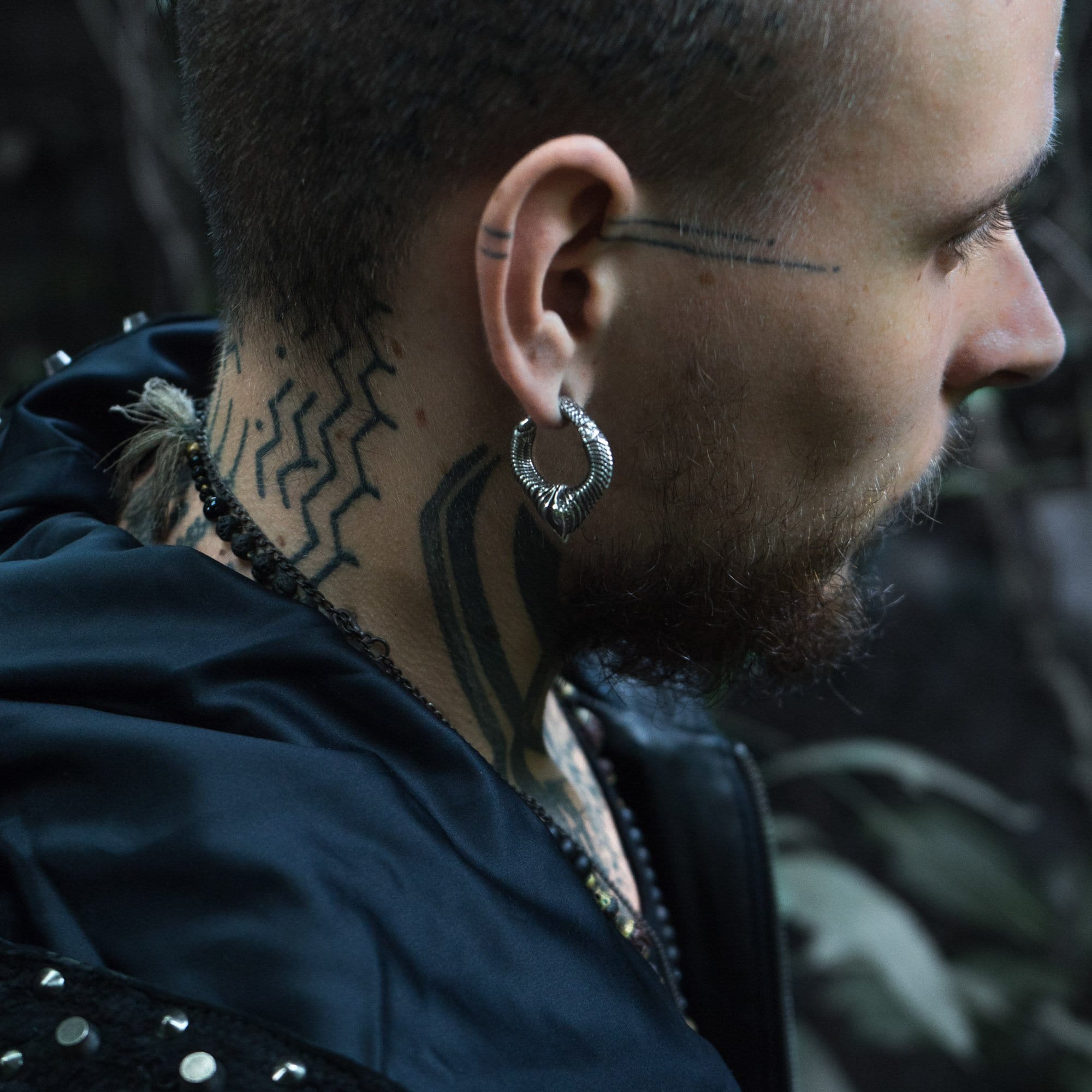 In profile, a person with a shaved head and tattoos on their neck and scalp is depicted. They are wearing the MARVDASHT Small Biomechanical Clicker Ear Weights in Silver Brass by Kaya Movement, a large, textured hoop earring, and a beaded necklace reminiscent of post-apocalyptic body jewelry. The individual is clad in a dark jacket adorned with metal embellishments against a blurred and dark background.